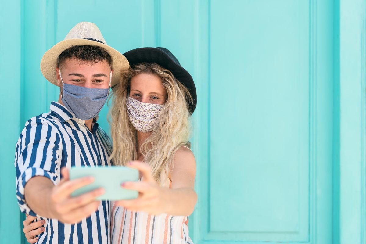 Young couple wearing face mask taking selfie with mobile smartphone on vacation - People having fun traveling again during corona virus outbreak - Love relationship and technology concept